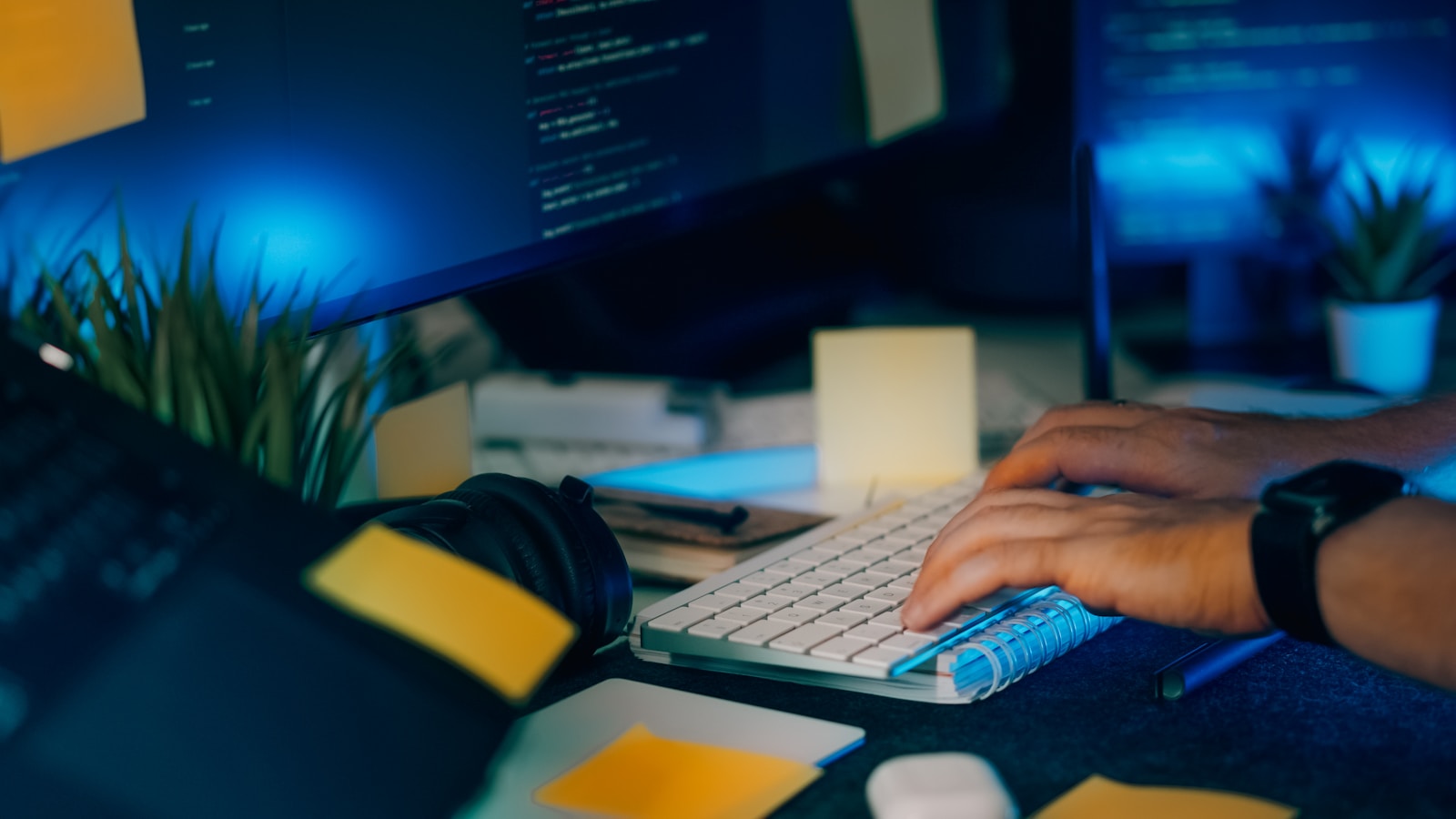 Hands typing on a keyboard with glowing computer screens in a dark home office